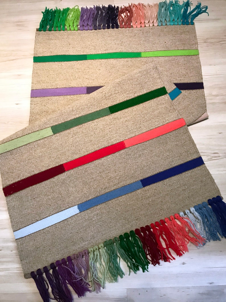 Puede incluir: Dos alfombras tejidas con flecos coloridos. La alfombra superior tiene rayas verdes, moradas y verdes. La alfombra inferior tiene rayas verdes, rojas, azules y blancas.