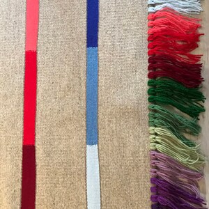 Puede incluir: Una alfombra tejida con fondo beige y dos rayas verticales. Las rayas son rojas, azules y blancas. La alfombra tiene un borde de flecos con borlas multicolores.