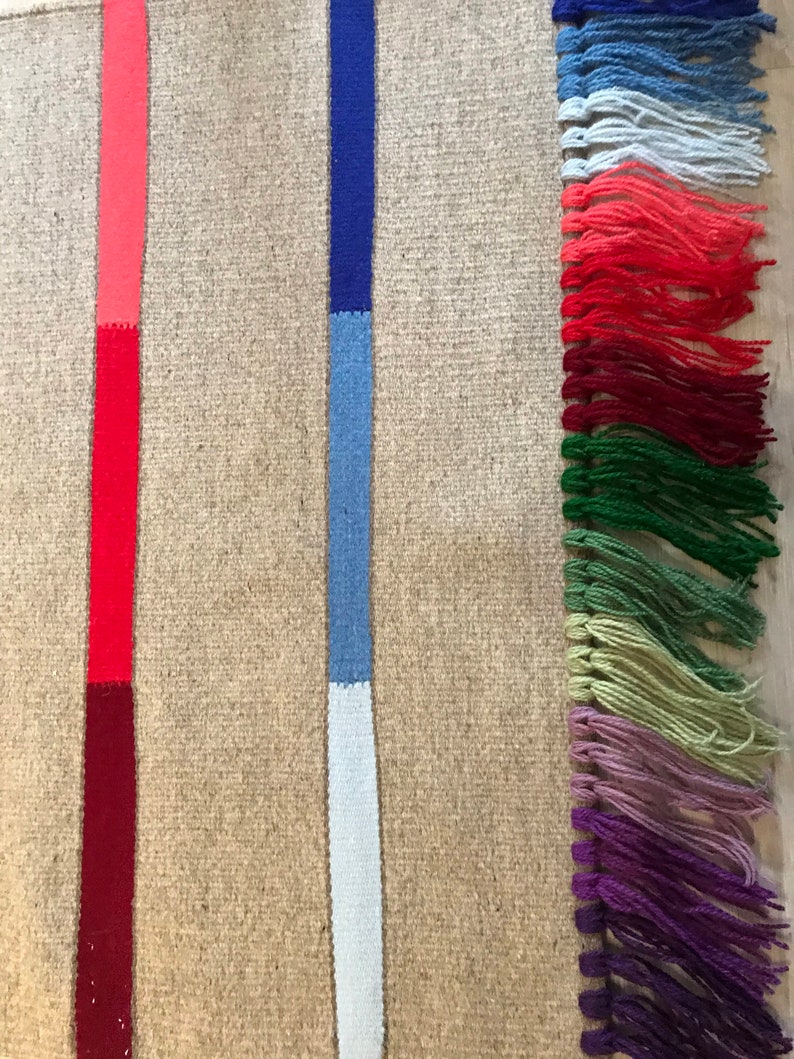 Puede incluir: Una alfombra tejida con fondo beige y dos rayas verticales. Las rayas son rojas, azules y blancas. La alfombra tiene un borde de flecos con borlas multicolores.