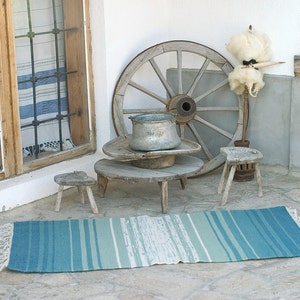 May include: A rustic porch setting with a wooden wheel, a metal pot, and three wooden stools. A blue and white striped rug is on the floor.