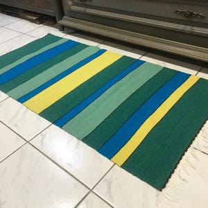 May include: A rectangular rug with a striped pattern in shades of green, blue, and yellow. The rug has a fringe on the short ends. The stripes run diagonally across the rug.
