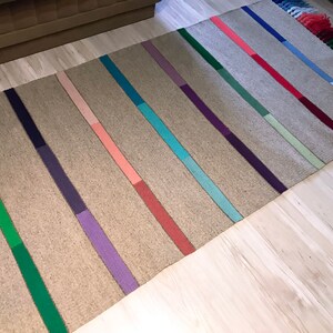 Puede incluir: Una alfombra tejida con un fondo marr&oacute;n claro y rayas de colores en tonos de verde, morado, rosa, azul y rojo. La alfombra tiene flecos en los extremos.