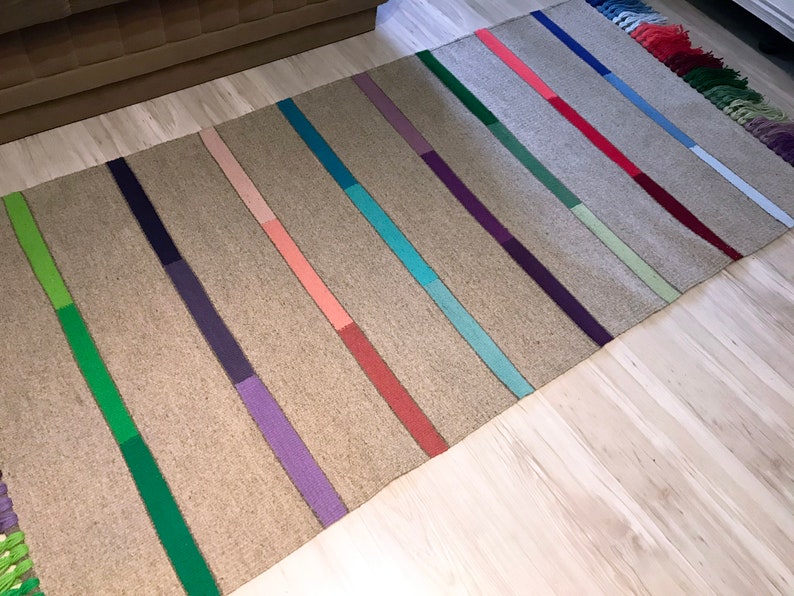 Puede incluir: Una alfombra tejida con un fondo marr&oacute;n claro y rayas de colores en tonos de verde, morado, rosa, azul y rojo. La alfombra tiene flecos en los extremos.