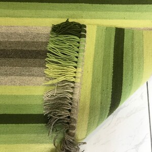 Può includere: Un tappeto tessuto a righe orizzontali nei toni del verde, marrone e beige. Il tappeto ha un bordo sfrangiato in varie tonalità di verde. Il tappeto è parzialmente arrotolato, rivelando il lato inferiore.