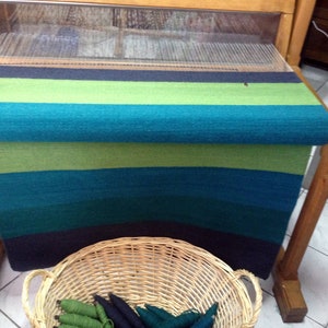 May include: A loom with a woven rug in shades of green, blue, and black. The rug is rolled up on the loom and there is a basket of yarn in the foreground.