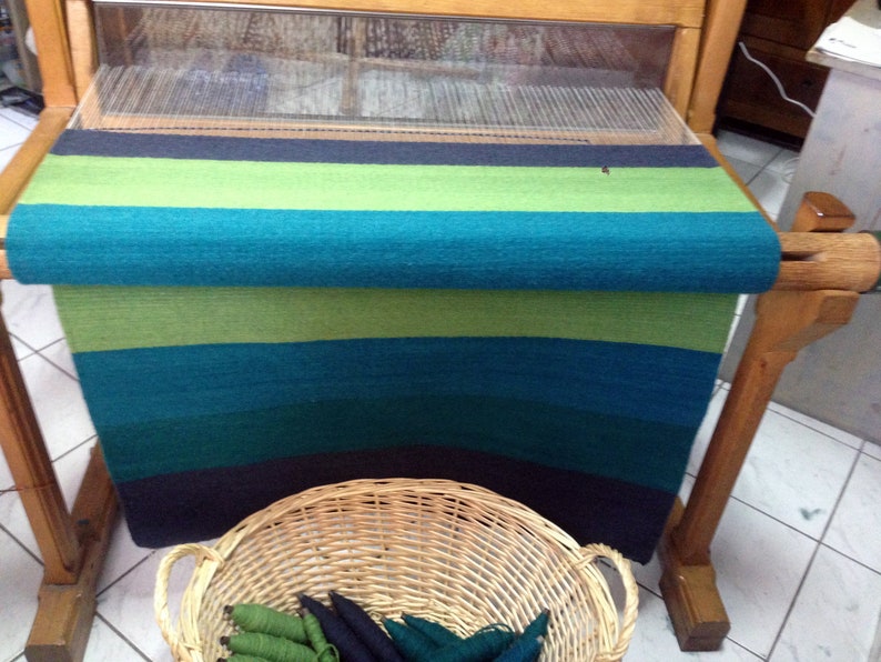 May include: A loom with a woven rug in shades of green, blue, and black. The rug is rolled up on the loom and there is a basket of yarn in the foreground.