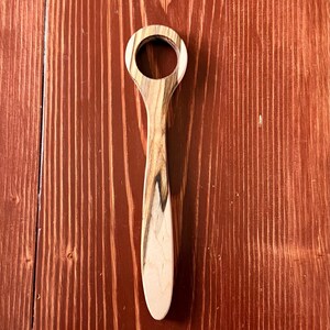 May include: A wooden bottle opener with a circular opening and a long, slender handle. The wood grain features a mix of light and dark brown tones. The opener is displayed on a reddish-brown wooden surface.