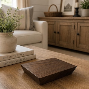 May include: A dark brown, square wooden tray with beveled edges sits on a light brown wooden coffee table. The tray is the focal point, with books and a vase of greenery nearby. The scene is set in a cozy living room.