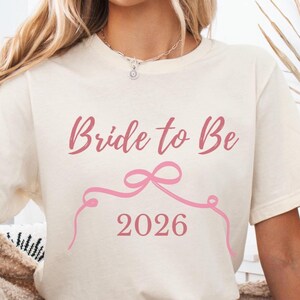 May include: Cream-colored t-shirt with the text "Bride to Be" in pink script, along with a pink bow and the year "2026". The shirt is a short-sleeved, casual style.