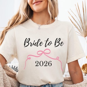 May include: Cream-colored t-shirt with the text "Bride to Be" in black script above a pink ribbon design and the year "2026" in black. The shirt is a casual style.