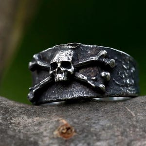 May include: A silver-toned ring featuring a skull and crossbones design. The ring has a wide band with a textured surface and a detailed skull at the center. The skull and bones are raised, creating a three-dimensional effect.