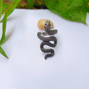 Op de afbeelding: Een zilverkleurige metalen ring in de vorm van een kronkelende slang met een textuur van schubben over het gehele oppervlak. De slangenkop bevindt zich aan de bovenzijde en het lichaam krult in een bocht naar beneden als ringband.