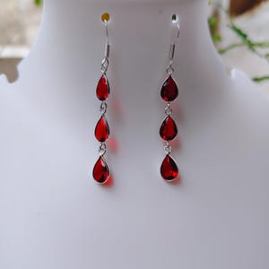 May include: A pair of long drop earrings featuring three faceted, teardrop-shaped red stones suspended in a vertical line. Each stone is encased in a silver-toned metal setting, connected by small silver rings, and attached to a silver hook ear wire.
