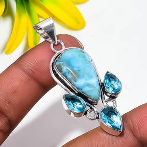 May include: A silver pendant featuring a large, pear-shaped, light blue Larimar stone at the center, accented by three smaller, faceted, pear-shaped blue topaz gemstones set in silver metalwork with a polished finish and a top bail.