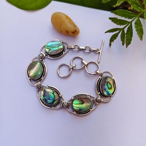 May include: A silver bracelet featuring oval-shaped abalone shells with iridescent green and blue hues. The bracelet has a toggle clasp and is set on a white surface. The abalone shells are framed by a decorative silver border.