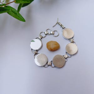 Puede incluir: Una pulsera de metal plateado con cinco bases circulares vacías conectadas por pequeñas anillas, terminada con un cierre de barra en T. Una piedra lisa de color beige está colocada en el centro de la disposición circular de la pulsera.