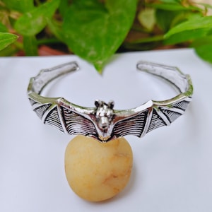 May include: A silver-toned metal cuff bracelet featuring a central bat motif with outstretched wings. The design includes detailed engraved lines on the wings and a sculpted bat head in the center, with an open back for adjustable wear.