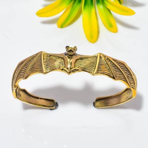 May include: A gold-toned metal cuff bracelet featuring a detailed bat design with outstretched wings. The bat is centered on the band, with its wings extending along the sides. The metal has a polished finish with textured wing details.