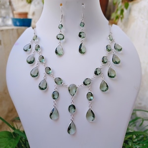 May include: A silver necklace and matching drop earring set featuring faceted, light green, oval and teardrop-shaped stones. The necklace has a multi-tiered design with dangling stones connected by silver links, while the earrings feature two stones.
