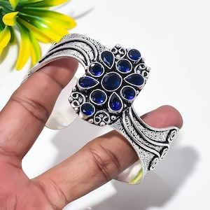 May include: A silver cuff bracelet featuring an intricate engraved pattern along the band and a central cluster of deep blue, faceted, pear-shaped and round gemstones arranged in a floral-inspired design with polished metal accents.
