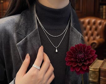 May include: A silver layered necklace featuring a delicate chain and a second chain with a smooth teardrop pendant. A silver band ring with a textured, repeating geometric pattern is worn on the finger. The jewelry is metallic and polished.