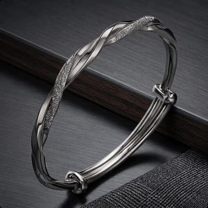 Puede incluir: Un brazalete de color plata oscura con un diseño trenzado. La banda alterna entre un acabado metálico liso y pulido y una superficie texturizada y brillante. El brazalete cuenta con un mecanismo de superposición ajustable en la base.
