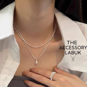 May include: A silver layered necklace with a teardrop pendant. The necklace is worn over a white collared shirt. A silver ring with a decorative design is on the finger. The text "THE ACCESSORY LABUK" is visible.