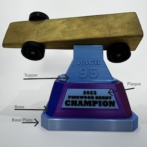 May include: A Pinewood Derby trophy featuring a wooden car with black wheels atop a blue, purple, and gray base. The base includes the text "PACK 95" and "2023 PINEWOOD DERBY CHAMPION".