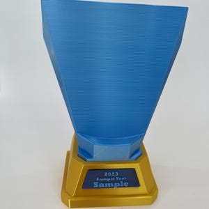 May include: A blue and gold trophy with the text "2023 Sample Text Sample". The trophy has a gold base and a blue, trapezoid-shaped top. The text is in white and blue.