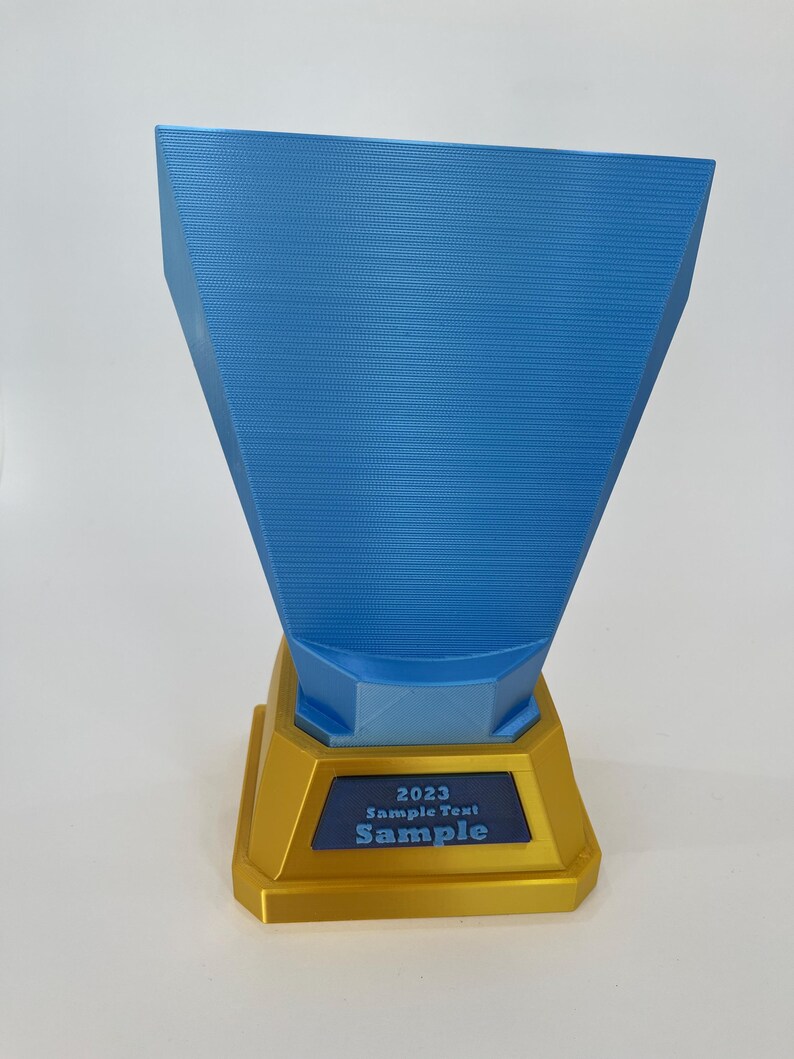 May include: A blue and gold trophy with the text "2023 Sample Text Sample". The trophy has a gold base and a blue, trapezoid-shaped top. The text is in white and blue.