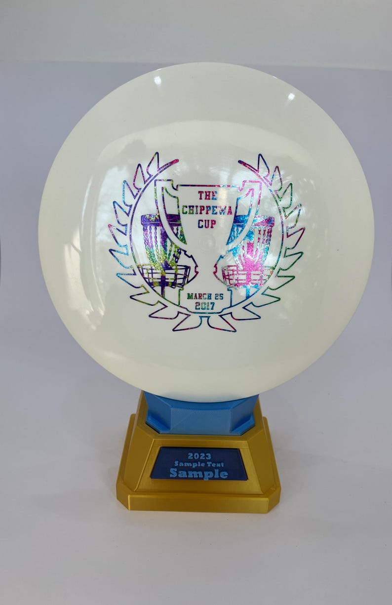 May include: A white disc golf trophy with a gold base and blue accents. The disc features a colorful graphic with the text "THE CHIPPEWA CUP" and "MARCH 25 2017". The base has the text "2023 Sample Text Sample".