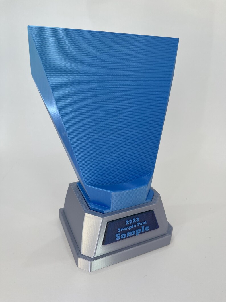 May include: A blue and gray trophy with the text "2023 Sample Text Sample". The trophy has a textured blue top section and a gray base. The text is in a dark blue rectangle.