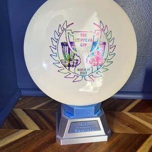 May include: A white disc golf disc with a colorful graphic of a trophy and text that reads "The Chippewa Cup" and "March 25 2017". The disc is displayed on a blue and silver stand with the text "2023 Sample Text Sample".