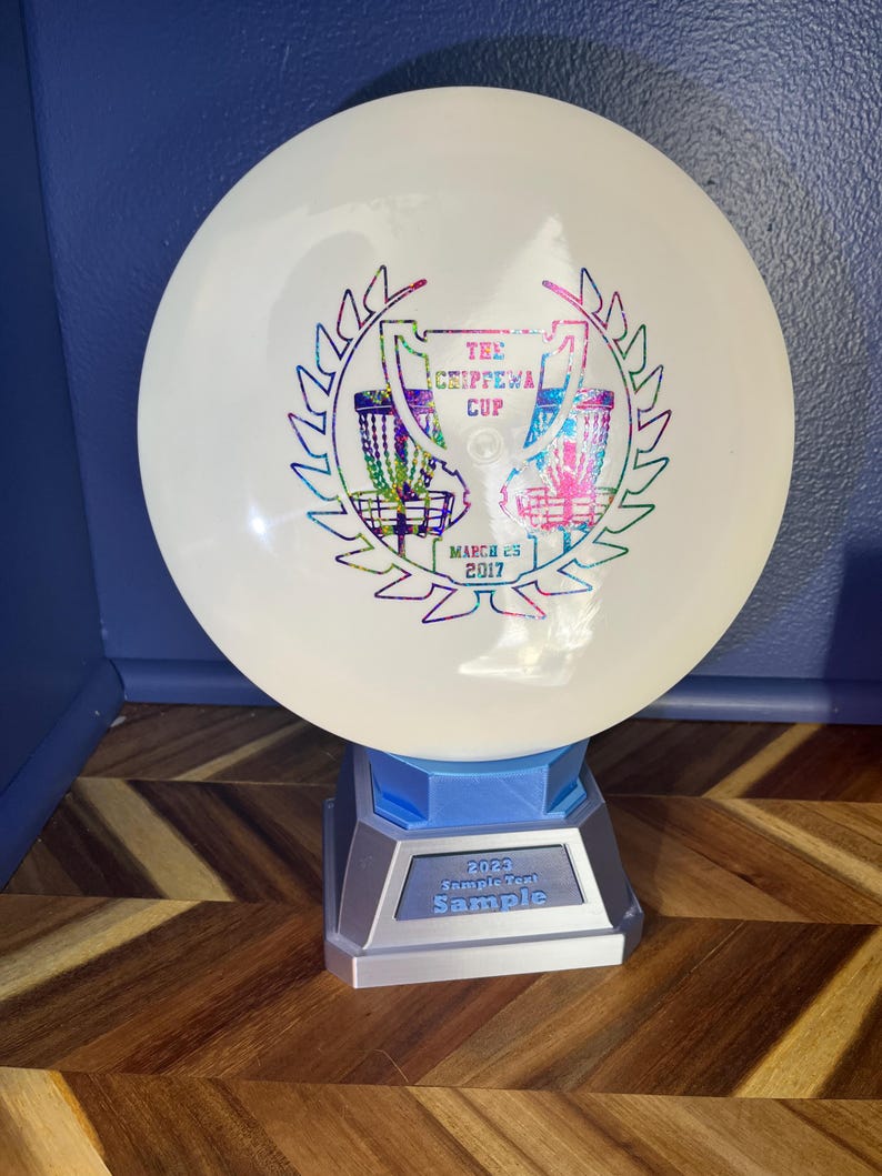 May include: A white disc golf disc with a colorful graphic of a trophy and text that reads "The Chippewa Cup" and "March 25 2017". The disc is displayed on a blue and silver stand with the text "2023 Sample Text Sample".