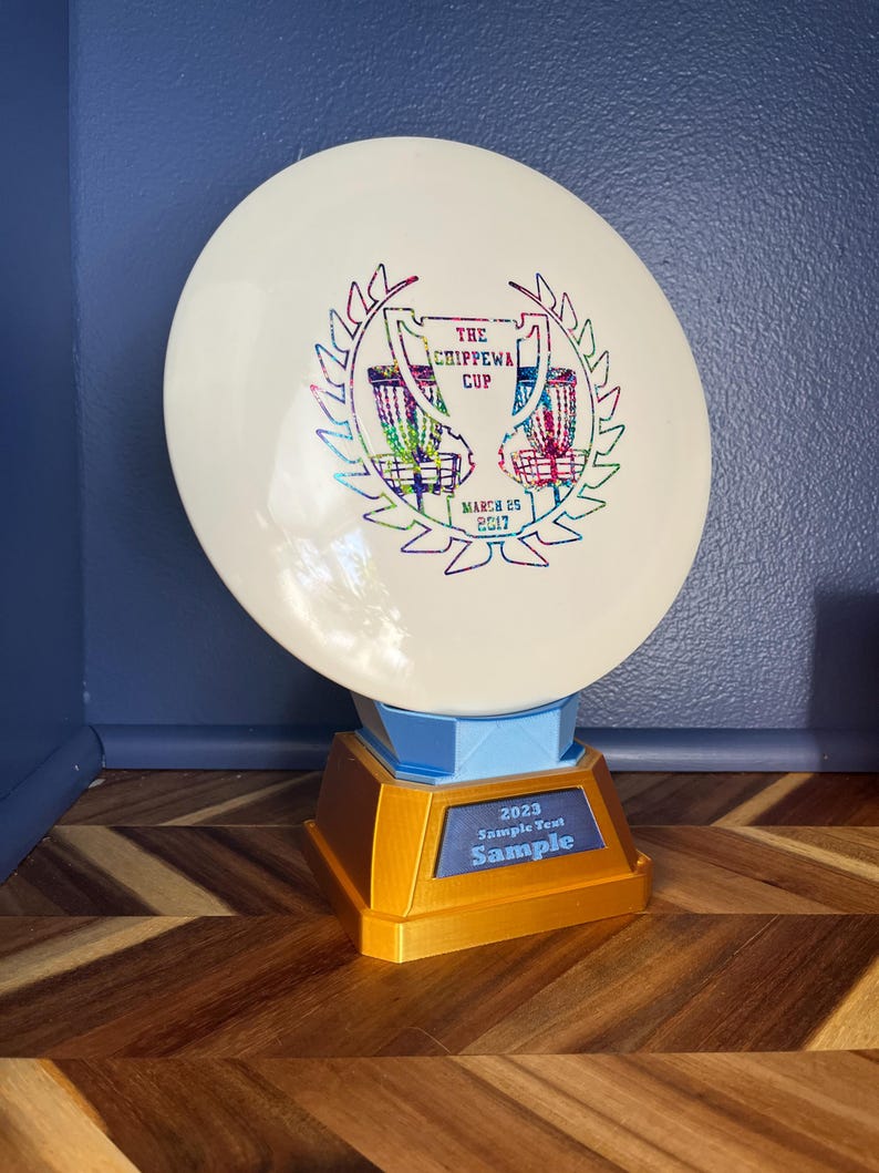 May include: A white disc golf trophy with a gold and blue base. The disc features the text "The Chippewa Cup" and "March 25 2017". The base has the text "2023 Sample Text Sample".