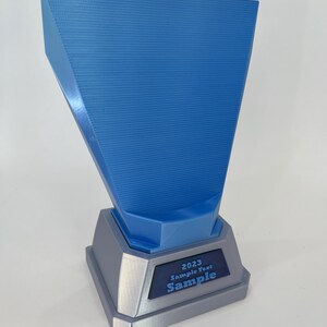 May include: A blue and gray trophy with the text "2023 Sample Text Sample". The trophy has a textured blue top section and a gray base. The text is in a dark blue rectangle.