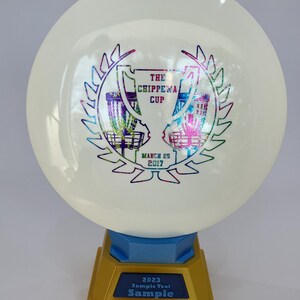 May include: A white disc golf disc with a colorful graphic of two baskets and text that reads "THE CHIPPEWA CUP MARCH 25 2017". The disc is mounted on a gold and blue trophy base with the text "2023 Sample Text Sample".