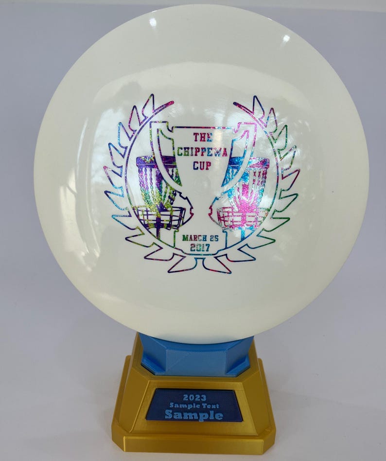 May include: A white disc golf disc with a colorful graphic of two baskets and text that reads "THE CHIPPEWA CUP MARCH 25 2017". The disc is mounted on a gold and blue trophy base with the text "2023 Sample Text Sample".