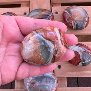 May include: Polished, oval-shaped stones in shades of red, brown, green, and cream. The stones have a smooth, glossy finish and are held in a hand. The background is a wooden surface. These stones are ideal for decorative purposes.