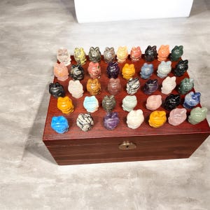 May include: A collection of small, carved frog figurines in various colors and materials, displayed on a wooden box. The frogs are in shades of blue, yellow, pink, green, and white, with some showing unique patterns and textures. The box is a dark brown color.