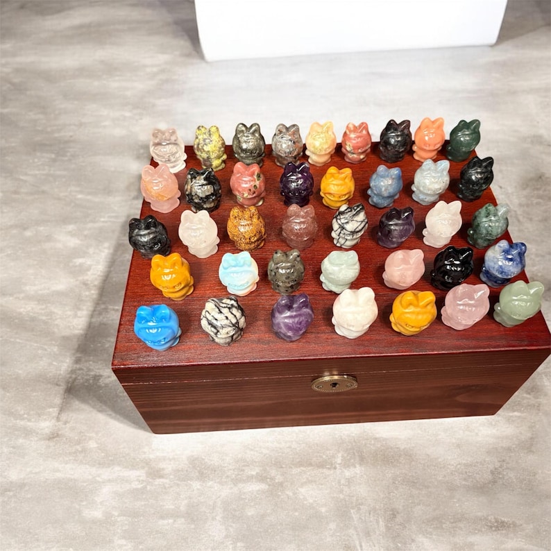 May include: A collection of small, carved frog figurines in various colors and materials, displayed on a wooden box. The frogs are in shades of blue, yellow, pink, green, and white, with some showing unique patterns and textures. The box is a dark brown color.