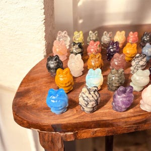 May include: A collection of small, carved cat figurines in various colors and materials, including pink, black, orange, and blue. The figurines are arranged on a wooden surface, showcasing their detailed features and unique textures.