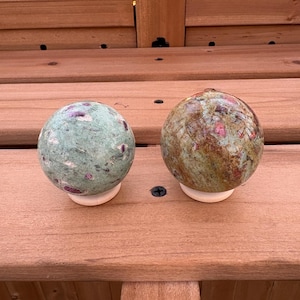 Peut inclure: Deux sphères décoratives en pierre, l'une verte avec des inclusions rouges, l'autre marron et verte avec des accents rouges. Chaque sphère repose sur une petite base circulaire blanche. Les sphères sont exposées sur une surface en bois.