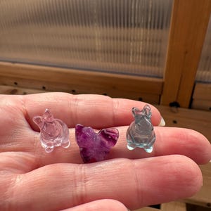 May include: Three small, carved crystal figurines. Two are bunny-shaped, one clear and one blue-tinted. The third is a heart shape in a deep purple color. The figurines are held in a hand, with a blurred background.