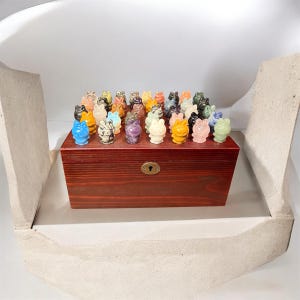 May include: A collection of small, colorful cat figurines displayed on a dark wooden box. The figurines are made of various materials and colors, including blue, yellow, pink, and green. The box has a keyhole and is set inside a white container.