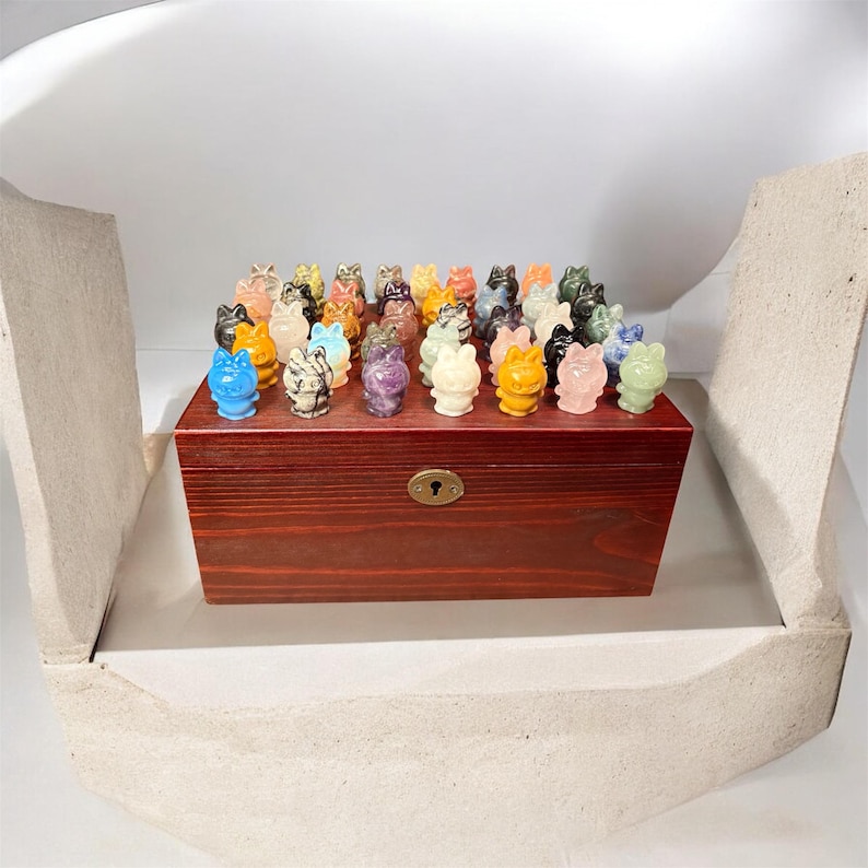 May include: A collection of small, colorful cat figurines displayed on a dark wooden box. The figurines are made of various materials and colors, including blue, yellow, pink, and green. The box has a keyhole and is set inside a white container.