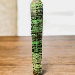 Op de afbeelding: Een rechthoekig kristal met een puntige top. Het kristal heeft een levendig groen en zwart gestreept patroon, met roze tinten aan de bovenkant. Het staat rechtop op een houten oppervlak.