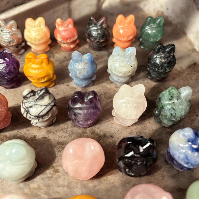 May include: A collection of small, carved gemstone frog figurines in various colors, including orange, green, black, and purple. The frogs are arranged closely together, showcasing the variety of materials and hues.
