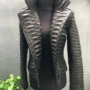 May include: A black, textured jacket with a unique, scale-like pattern. The jacket features a high, structured collar and a full-length zipper. The sleeves are long, and the jacket is displayed on a black mannequin.