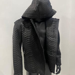 May include: A black hooded jacket with a textured, scale-like pattern. The jacket has a zipper closure and a hood. The jacket is displayed on a black mannequin. The jacket is made of a material that resembles snakeskin.